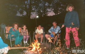 Kilka osób siedzi na krzesłach biwakowych przy ognisku. Fotografia wykonana wieczorem