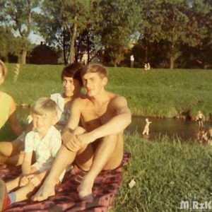 Fotografia kolorowa. Rodzina siedzi na kocu na trawie.